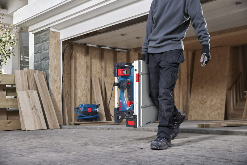 Eine Person mit Sicherheitsausrüstung trägt auf einer Baustelle eine tragbare Tischkreissäge. Anwendungsbild Nahaufnahme